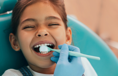 A young girl smiling in a dental chair while a dentist cleans her teeth with a dental tool.