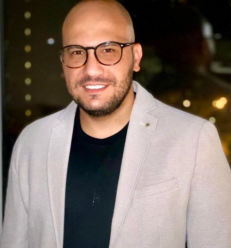 A smiling man with a shaved head and glasses, wearing a light gray blazer over a black shirt, stands in an illuminated setting.