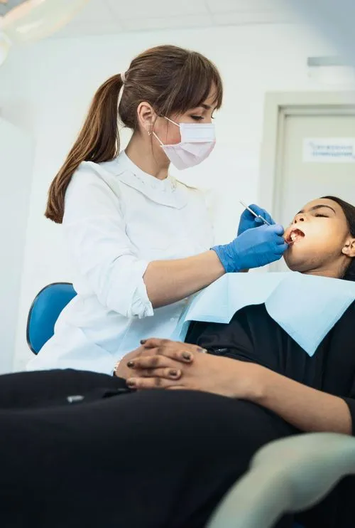 A dentist in a white coat and mask examines a patient's teeth in a dental treatment room.