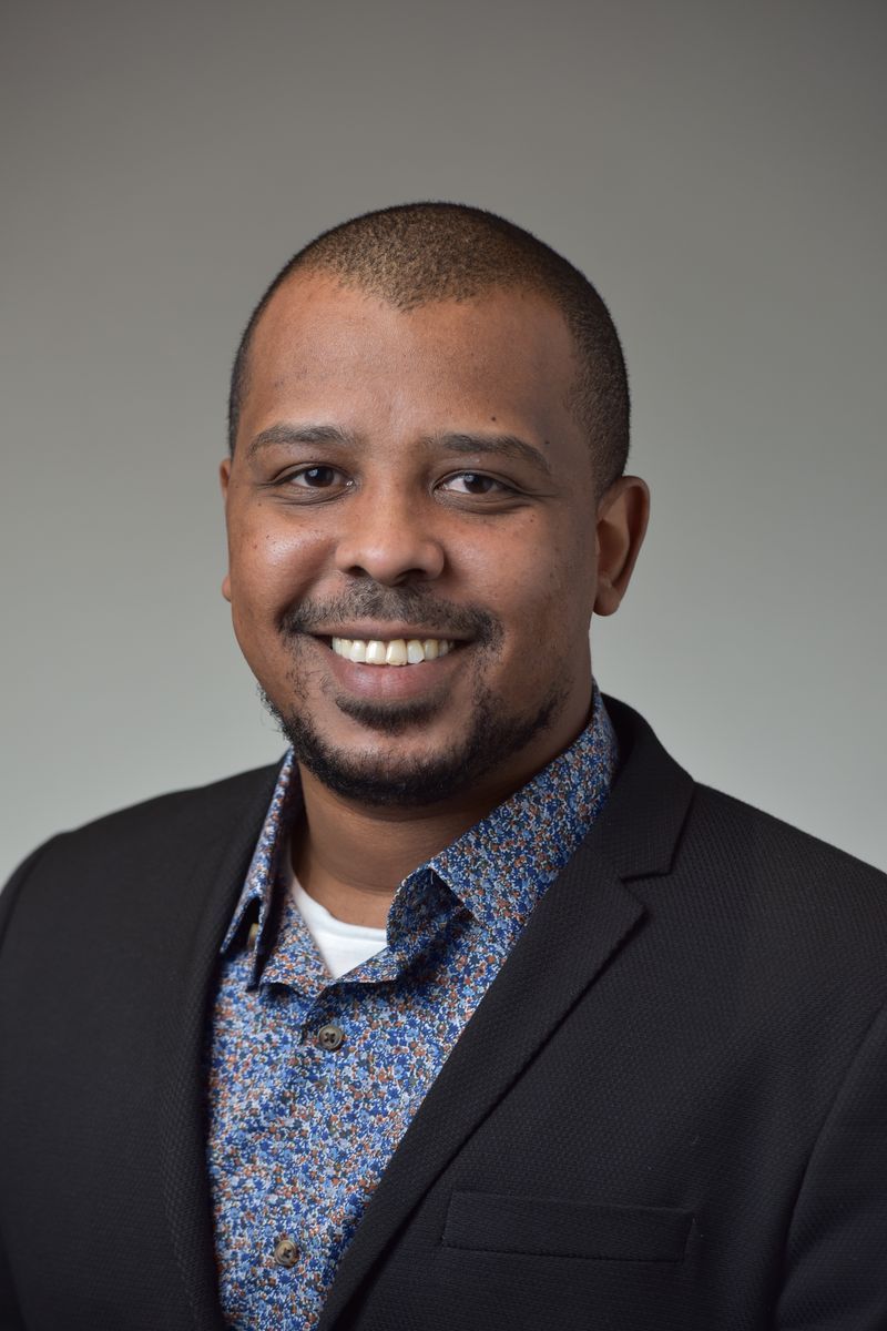 A smiling man with short hair wearing a black blazer over a floral shirt poses against a neutral background.