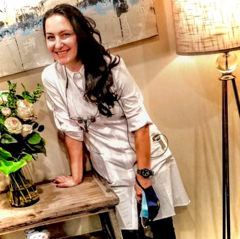 A dentist in a white coat smiles while leaning against a wooden table next to a vase of flowers and a lamp.