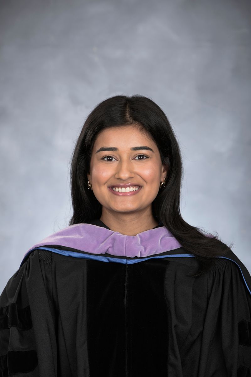 A smiling female dentist in graduation robes poses for a portrait against a gray backdrop.